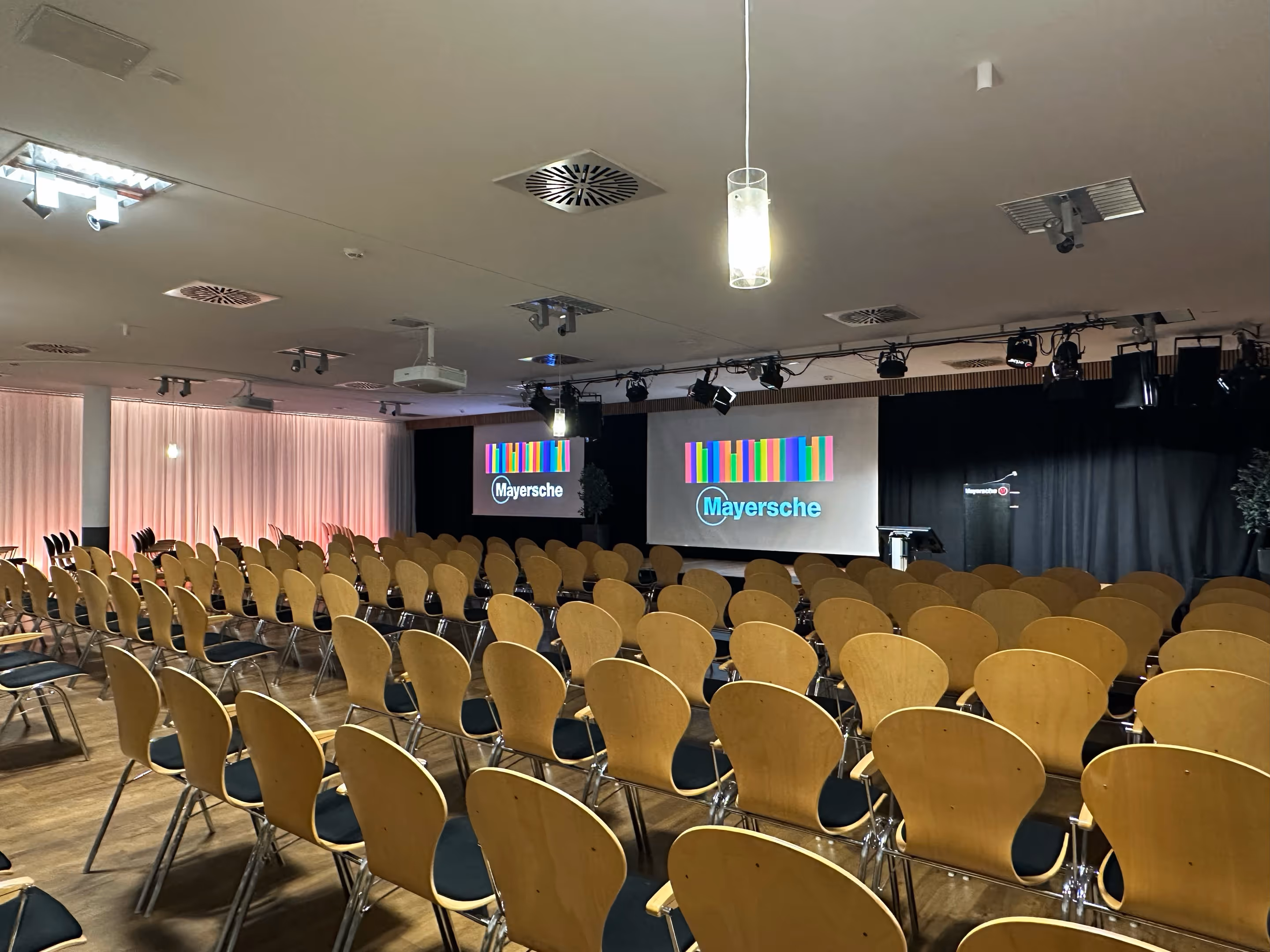 Empty conference room with rows of wooden chairs facing dual screens.