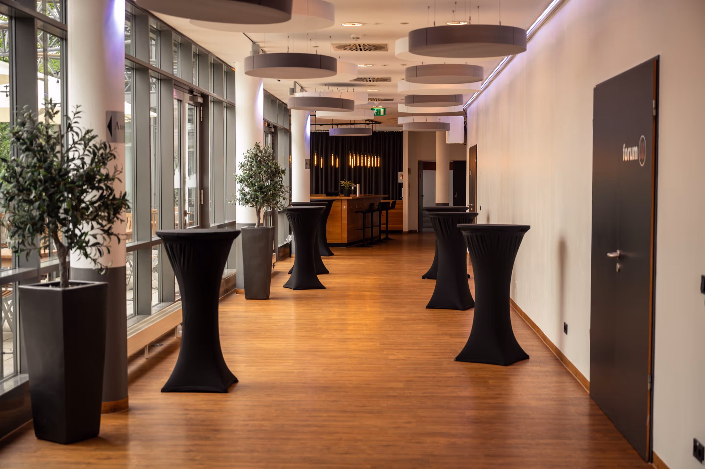 Modern event space hallway with wooden floor, tall cocktail tables covered in black cloth, large potted plants, and pendant lights above.