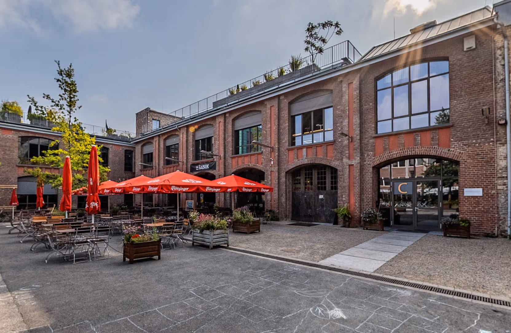 Outdoor seating area with red umbrellas and metal tables in front of a brick building with large arched windows and a sign reading La Fabrik.