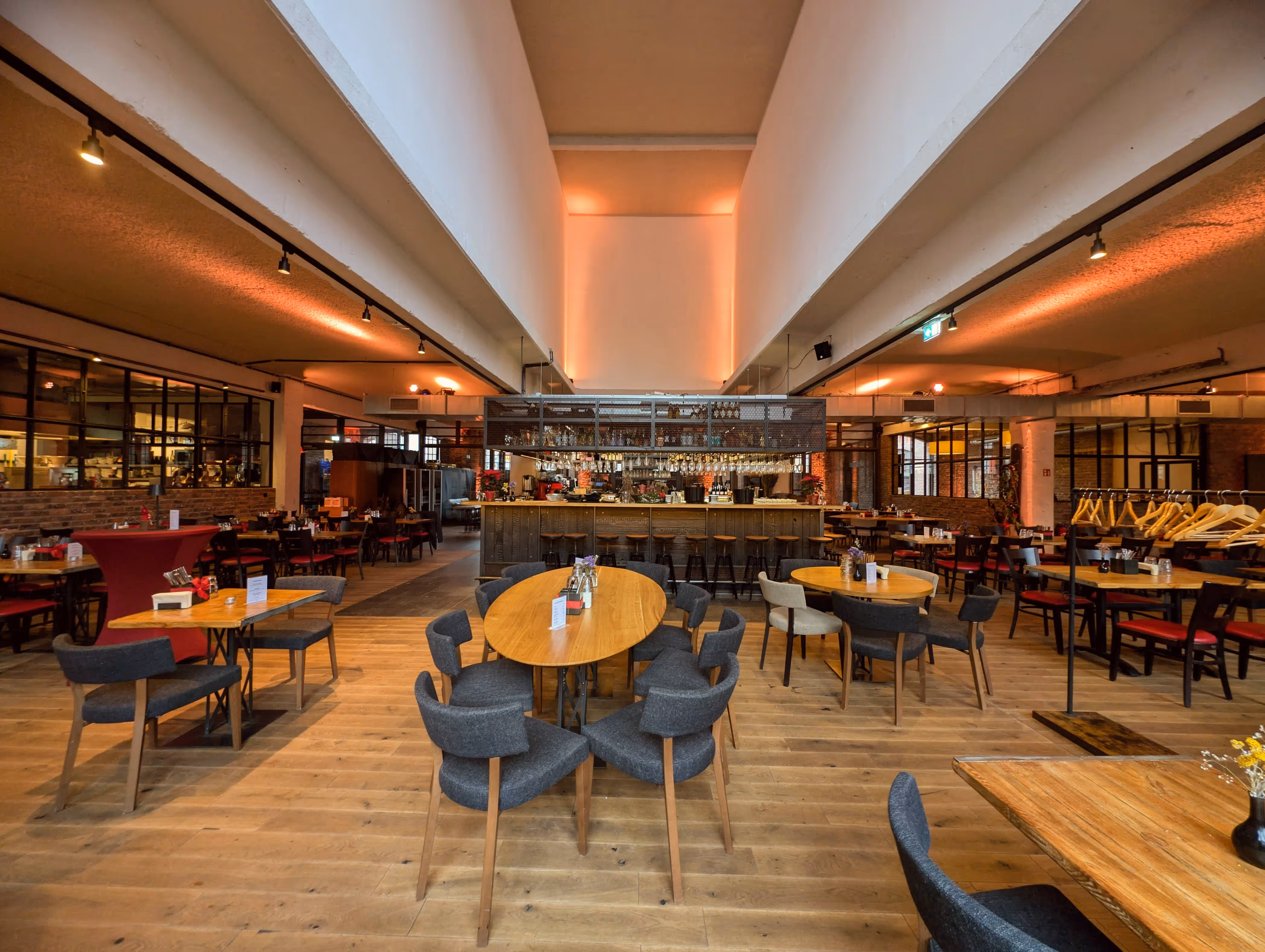 Spacious modern restaurant interior with wooden floors, various round and oval tables surrounded by gray and red cushioned chairs, and a central bar area with hanging glasses and warm ambient lighting.