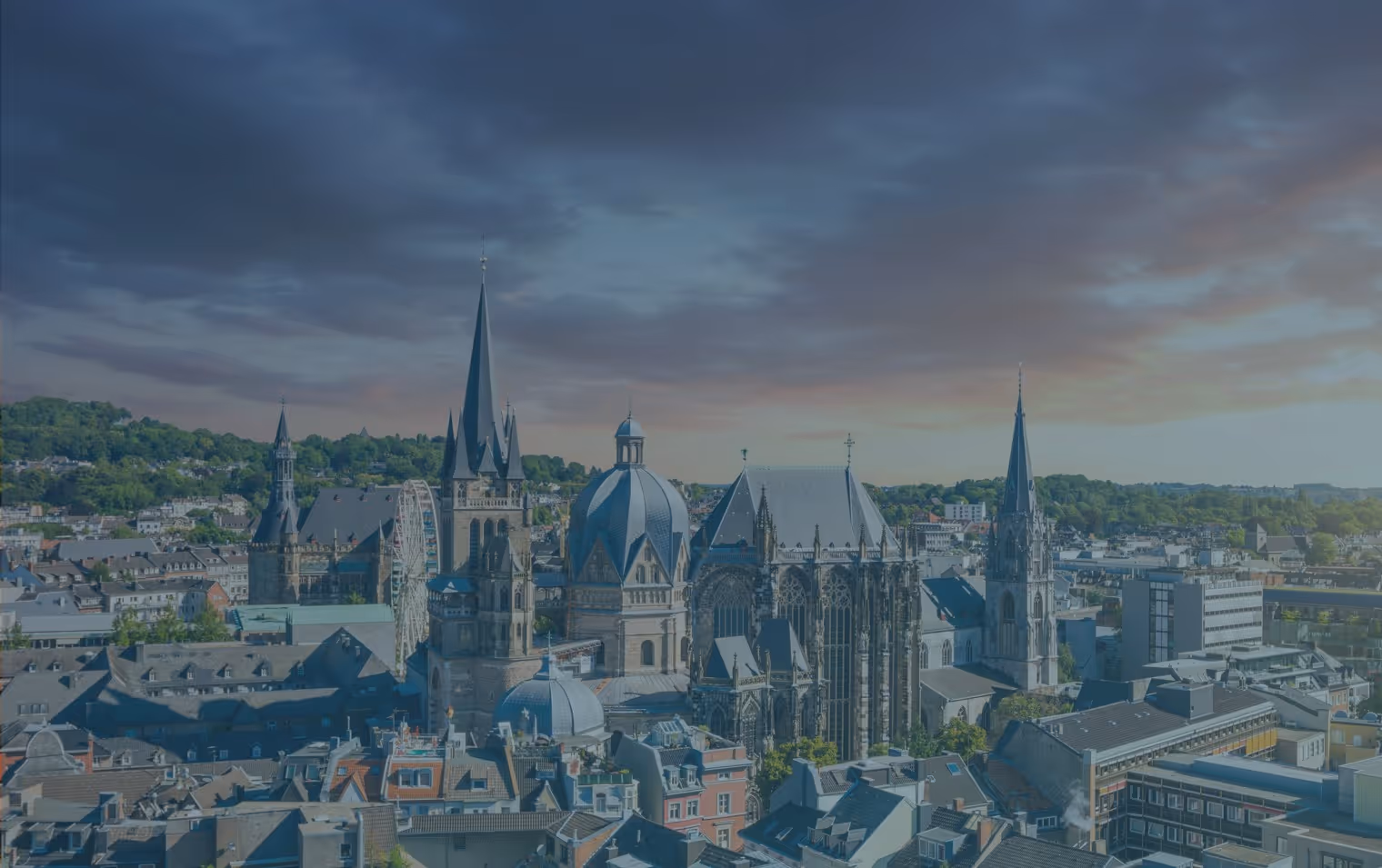 Cityscape of Aachen featuring historic Gothic and Baroque church buildings with prominent spires under a colorful sunset sky.