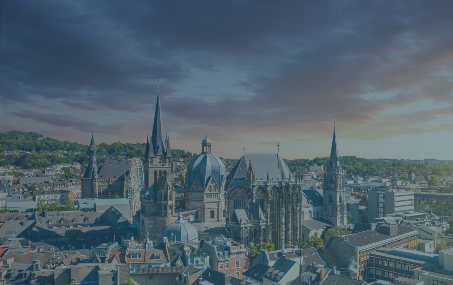 Cityscape of Aachen featuring historic Gothic and Baroque church buildings with prominent spires under a colorful sunset sky.