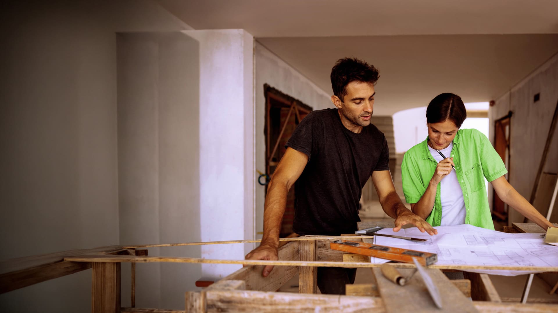 Deux personnes examinent un plan sur une table de travail en bois dans un chantier de construction.