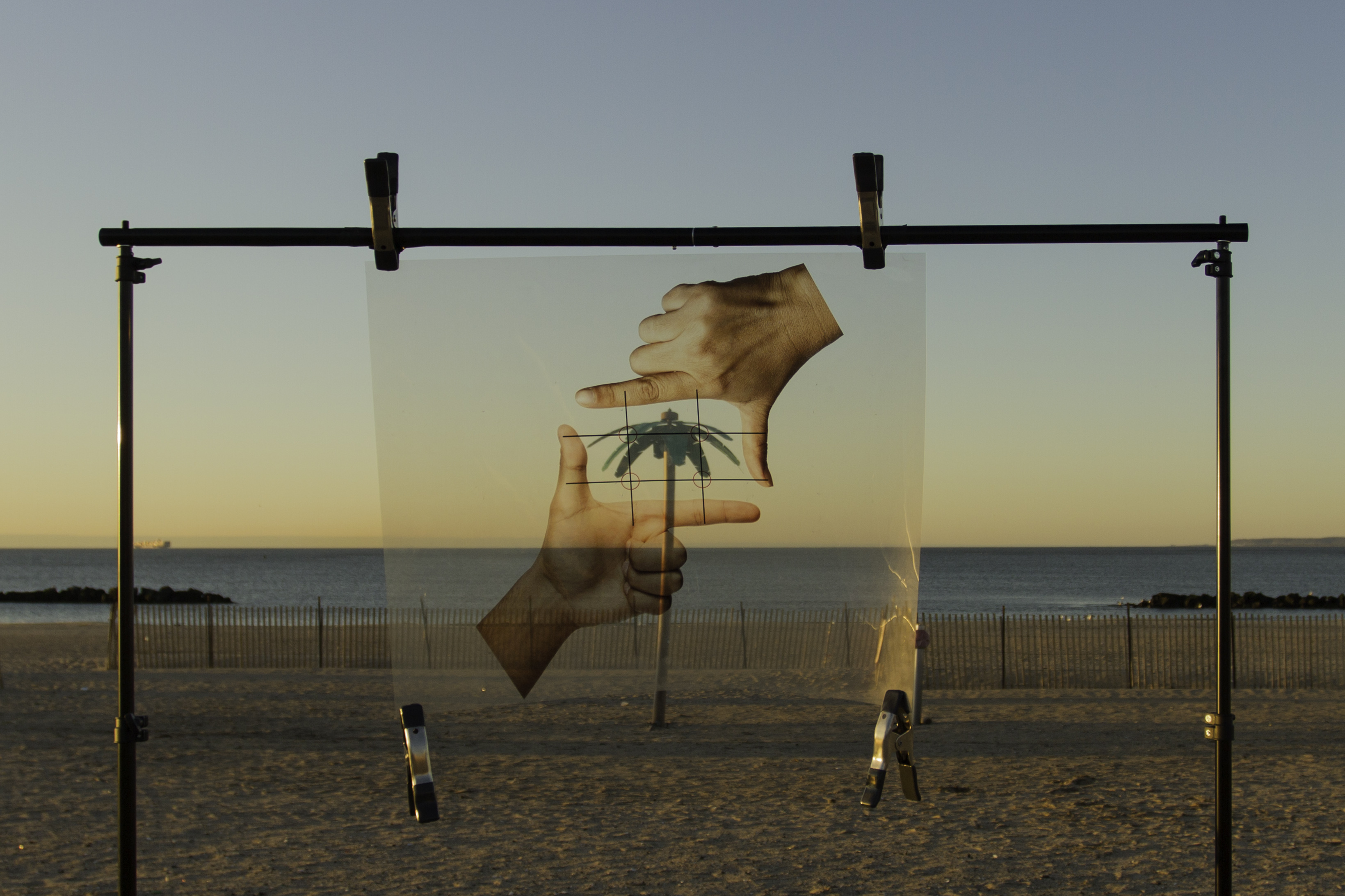 A fake palm tree on a beach visually masked with a clipped up transparency of two hands in a L shape framing the tree.