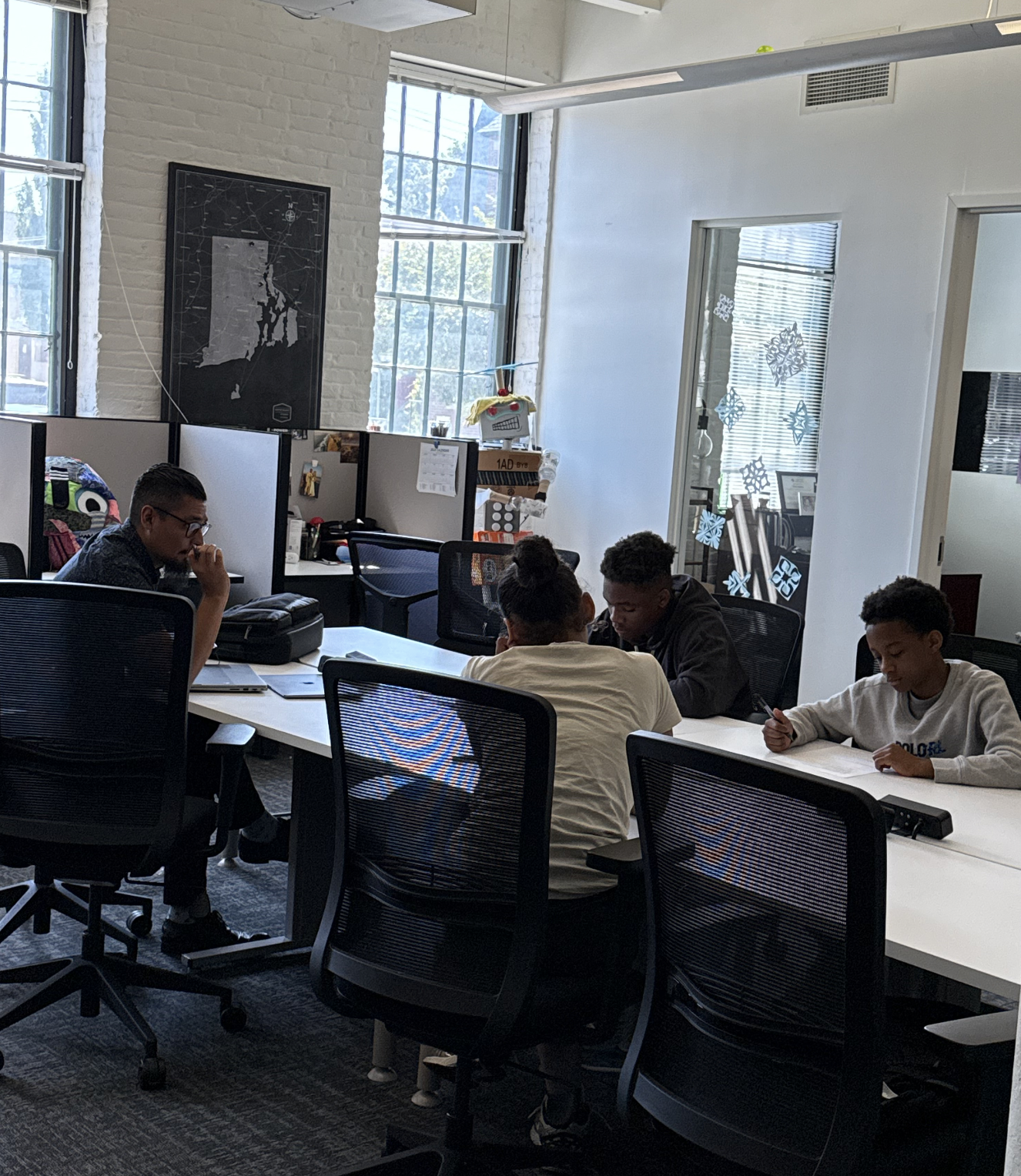 A group of teens and a mentor sit together at a long white table, working on assignments in a collaborative setting.