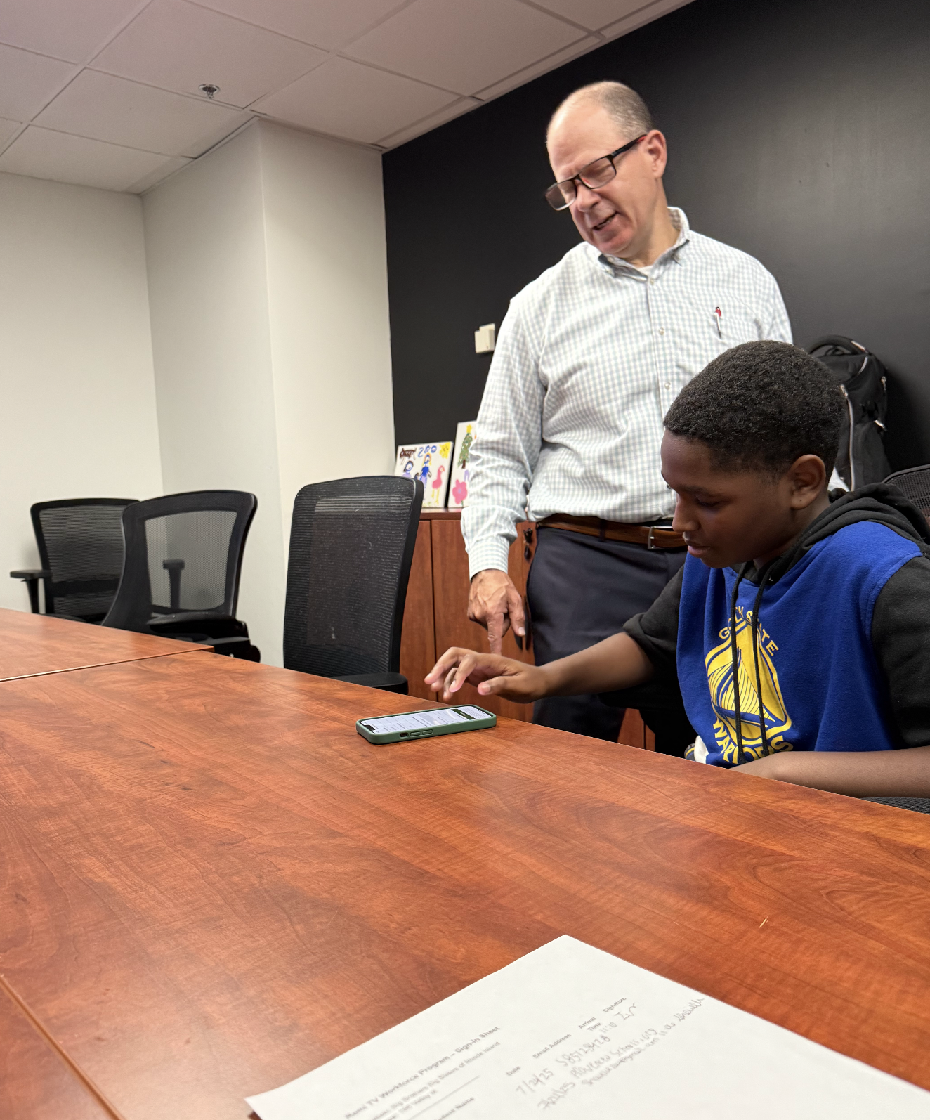A boy in a blue hoodie uses a smartphone while an adult mentor stands beside him, guiding him during a workshop.