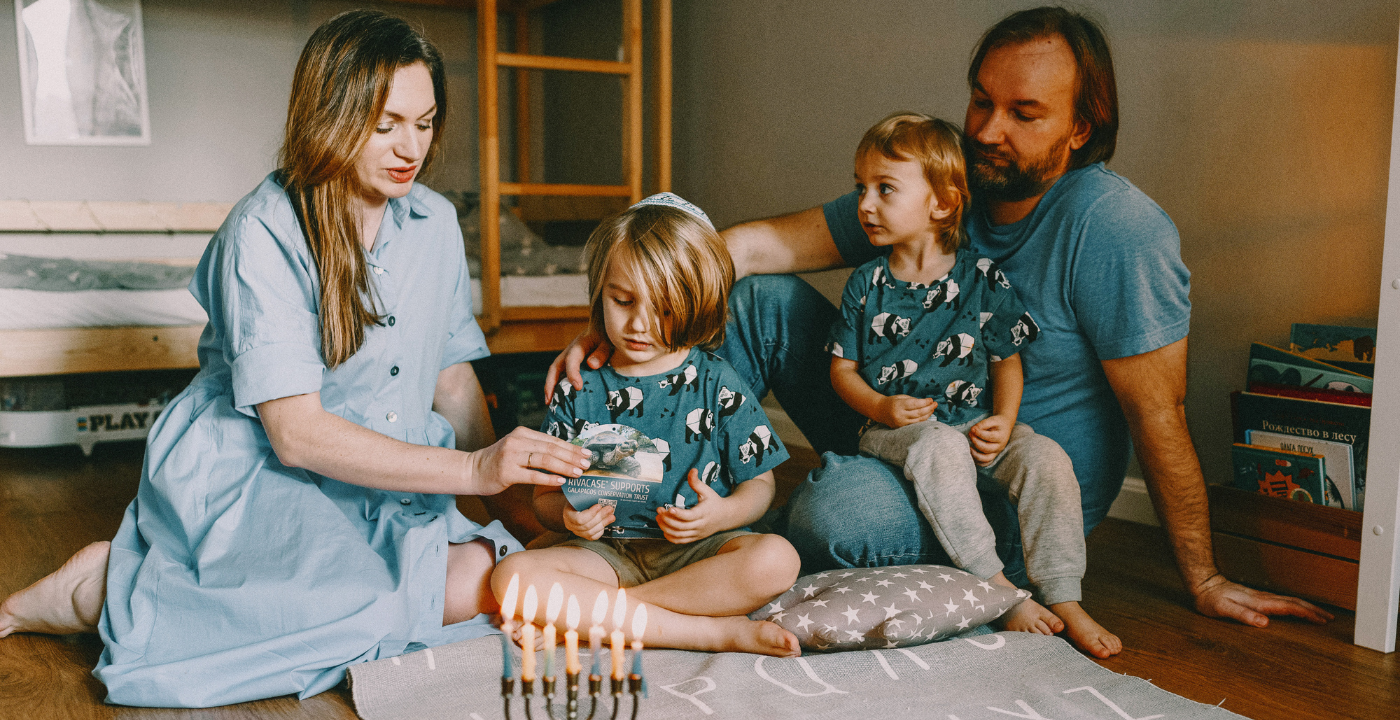 A mother and father sit on floor with two young children and light a menorah 