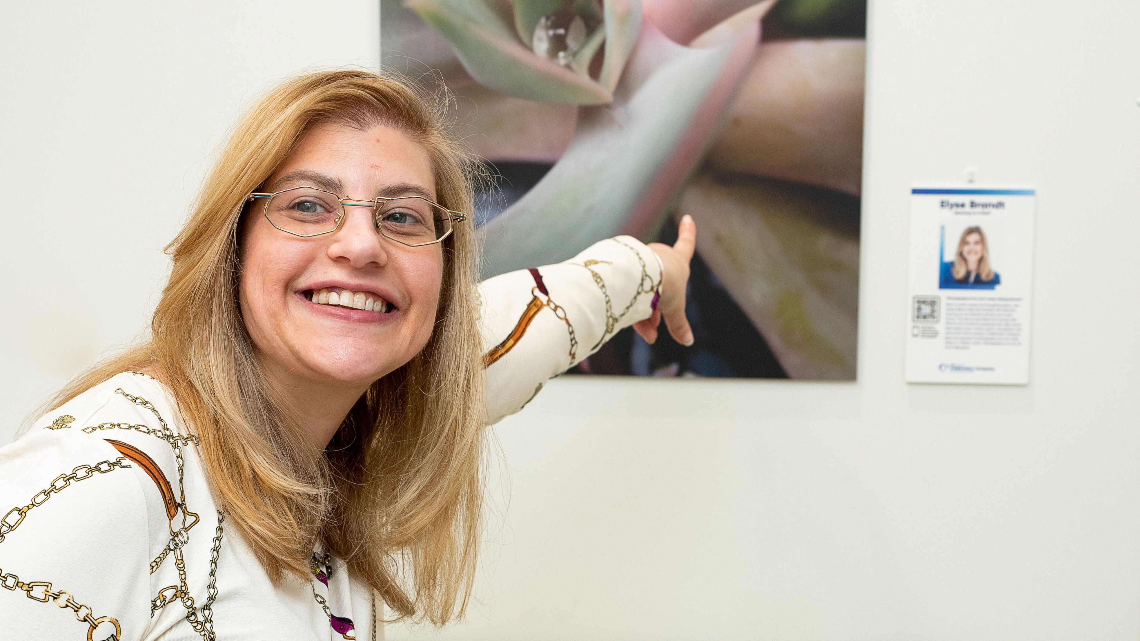 Celebrtion Company artist Elyse Brandt, smiling with long blonde hair and glasses, points proudly to her large photo of a succulent plant displayed on a wall behind her. A small plaque next to the photo features her portrait and name, identifying her as the artist.