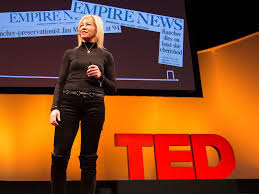 Judy MacDonald Johnston stands on stage delivering a TED Talk. She is dressed in black and holding a microphone. Behind her is a large screen displaying the title "EMPIRE NEWS" and a bold red TED logo on the stage.
