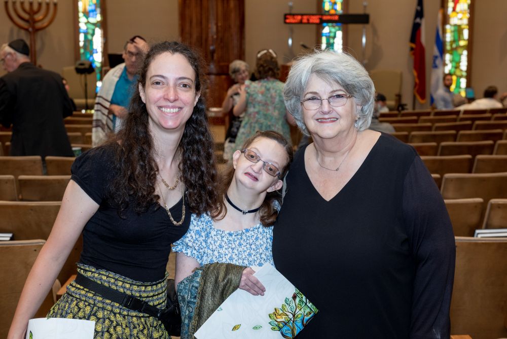 Three women smiling together inside a synagogue, with stained-glass windows and rows of seats in the background.