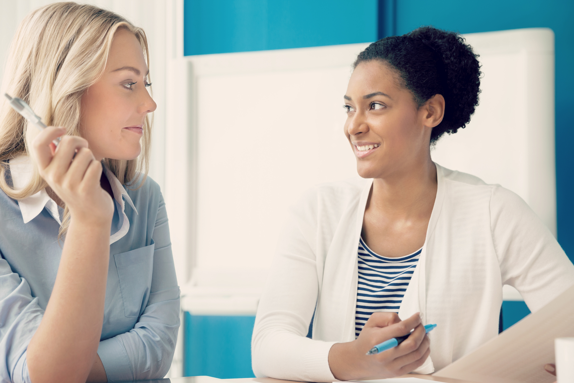 Two women sit across from each other in a bright office, smiling and engaged in conversation while holding pens, suggesting a friendly coaching or collaborative work session.