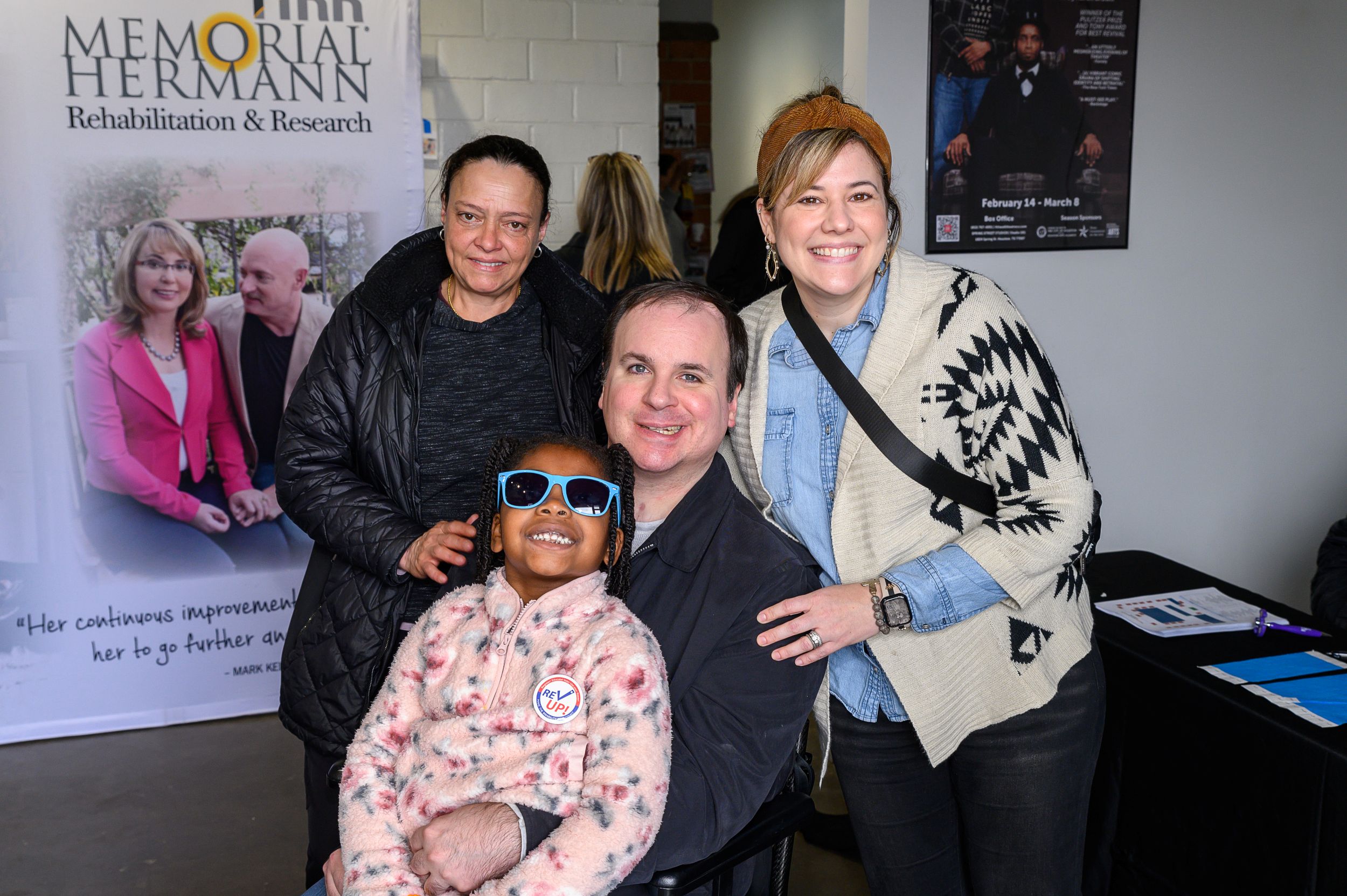 Four people smile together at an indoor community event. A man seated in a wheelchair holds a young child, while two women stand behind them with arms around their shoulders. A Memorial Hermann Rehabilitation & Research banner and event table are visible in the background.
