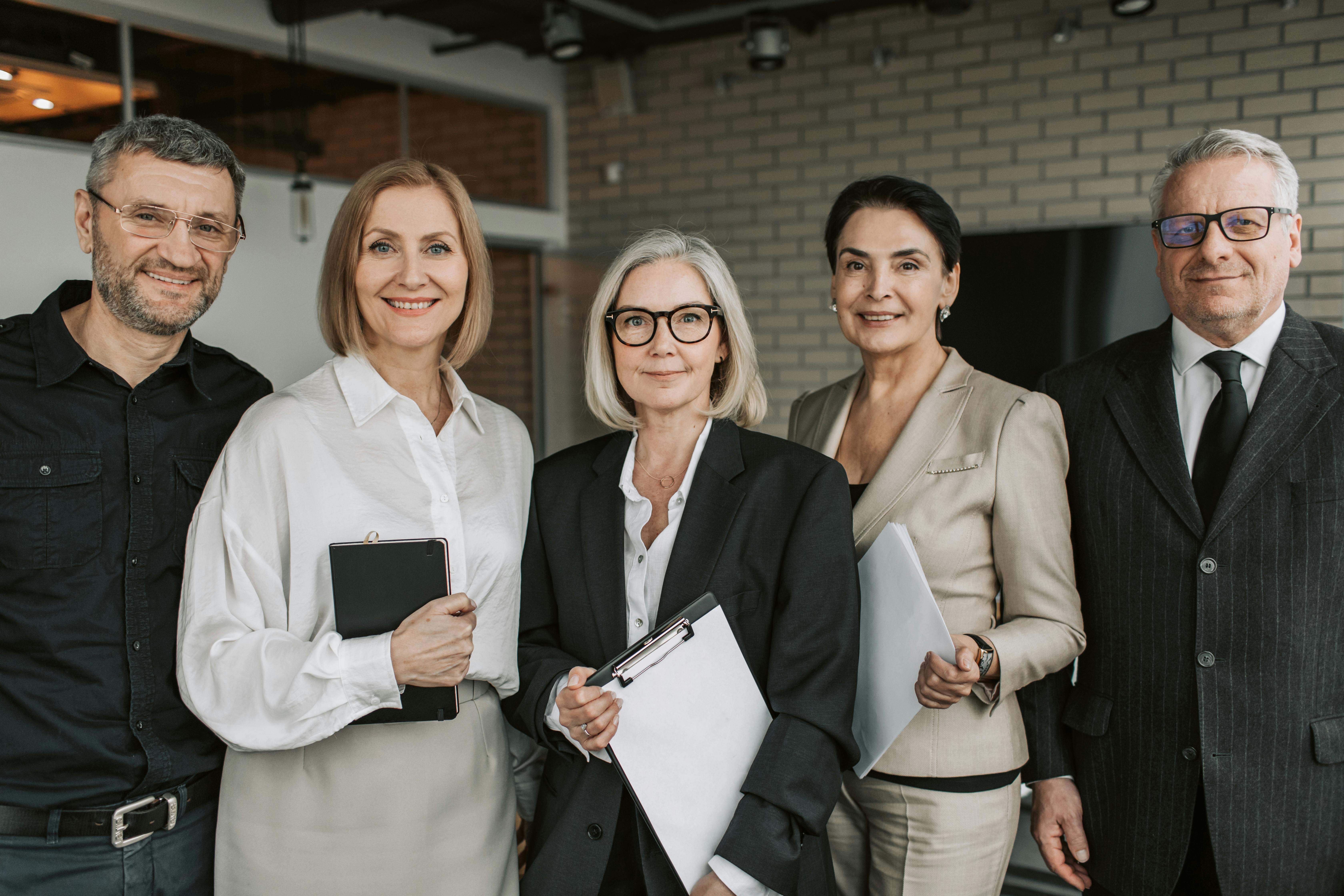 A diverse group of five professionals stand together in a modern office setting, smiling confidently at the camera.