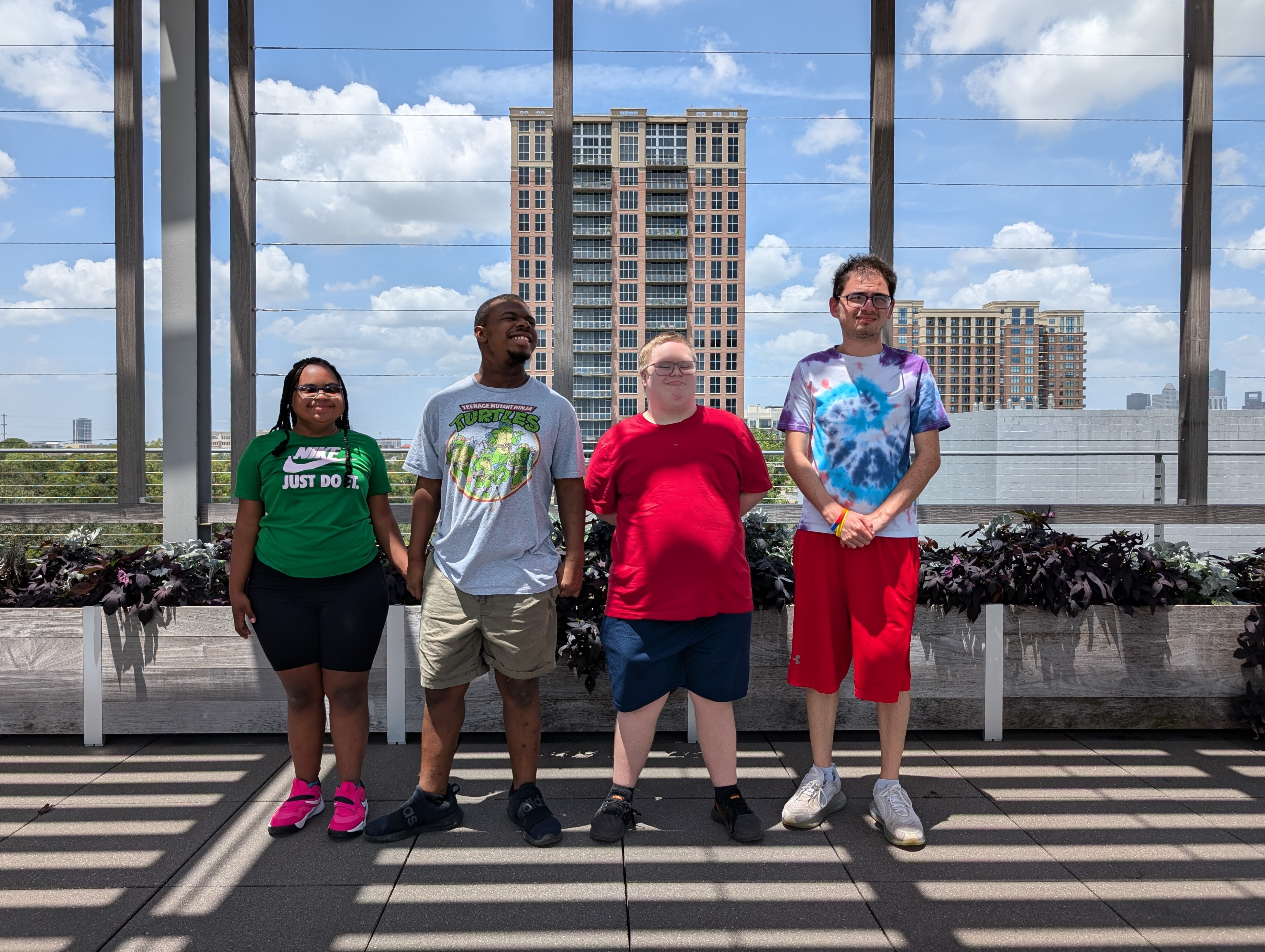 Four participants stand together on a sunny rooftop terrace with planters and a city skyline in the background.