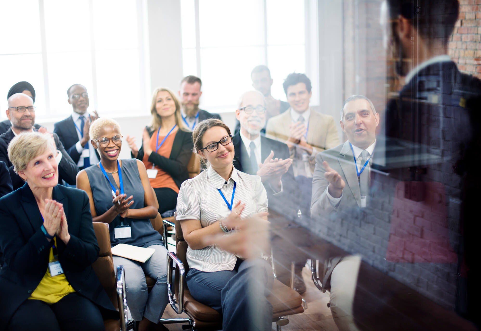 A diverse group of professionals seated in a bright meeting room applaud a speaker at the front of the room during a workplace training or presentation.