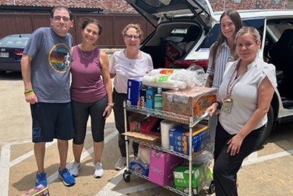 Five volunteers stand in a parking lot beside an open car trunk, smiling next to a rolling cart filled with donated supplies including paper goods, food items, and boxes.