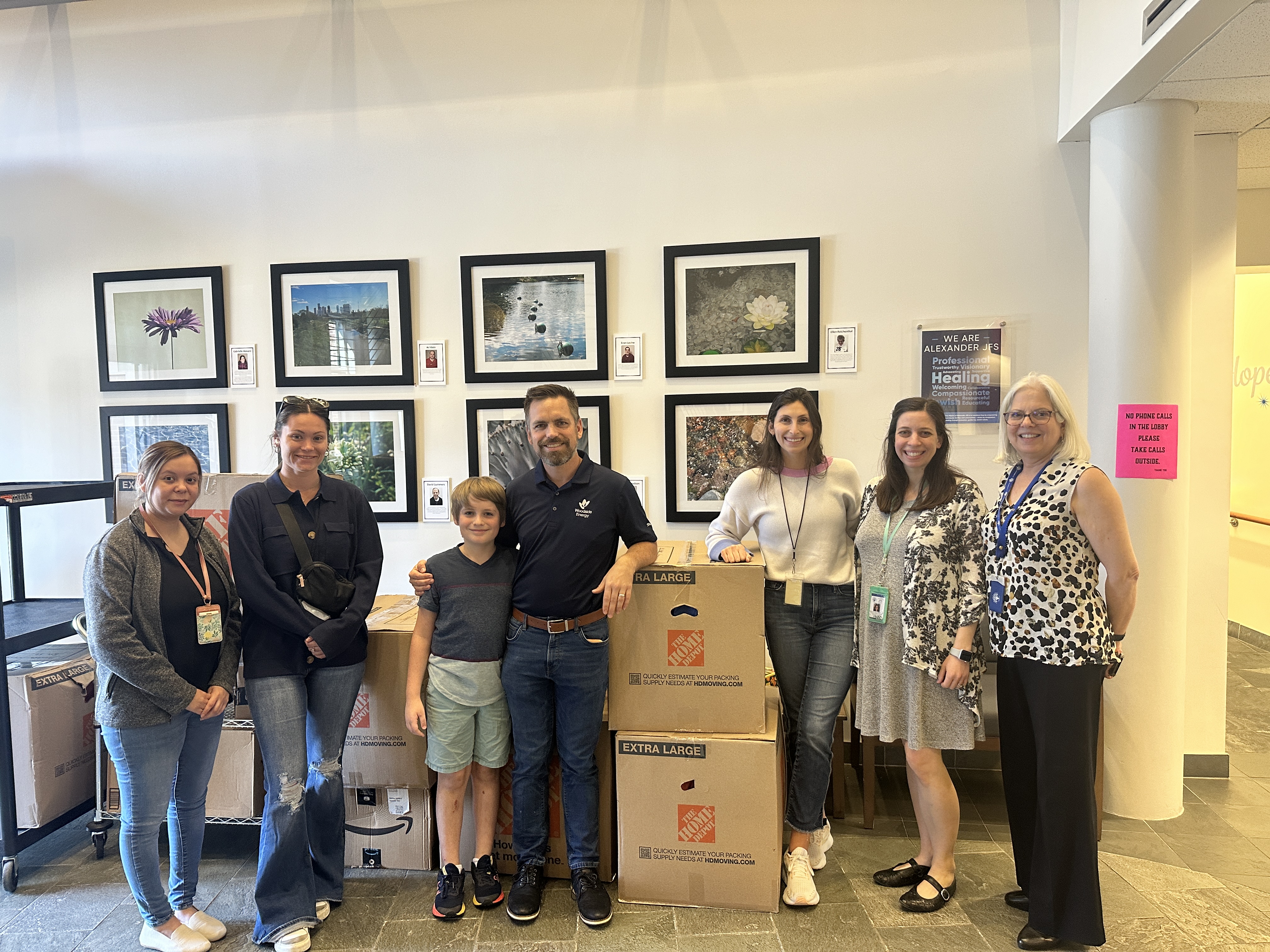 Five volunteers stand in a parking lot beside an open car trunk, smiling next to a rolling cart filled with donated supplies including paper goods, food items, and boxes.