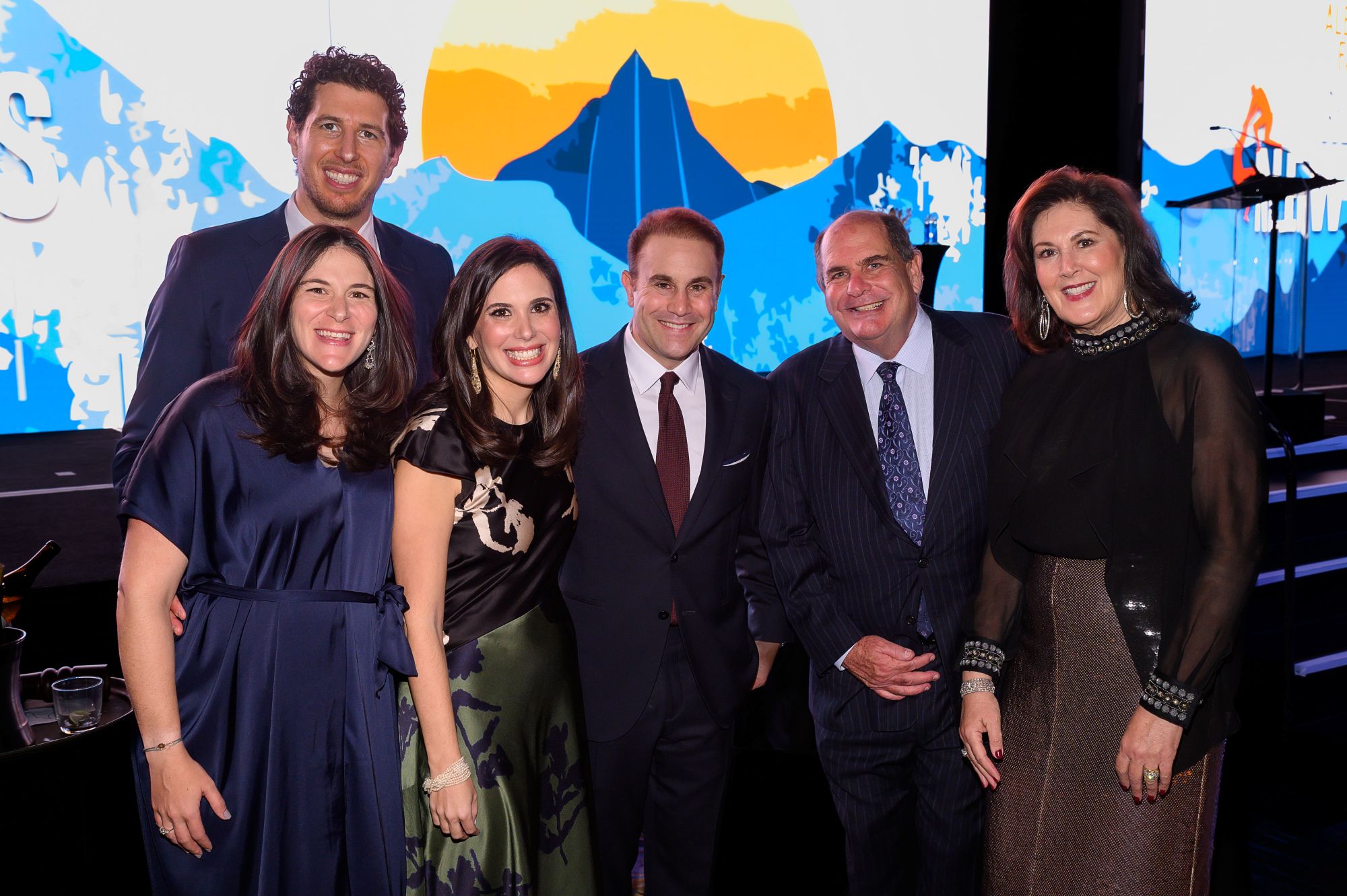 Six adults dressed in formal attire pose together at a gala event, smiling at the camera.