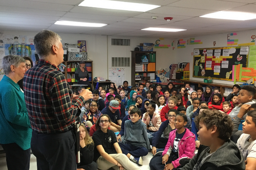 Two presenters speak to a large group of elementary school students seated in a classroom during an educational program or community outreach presentation.