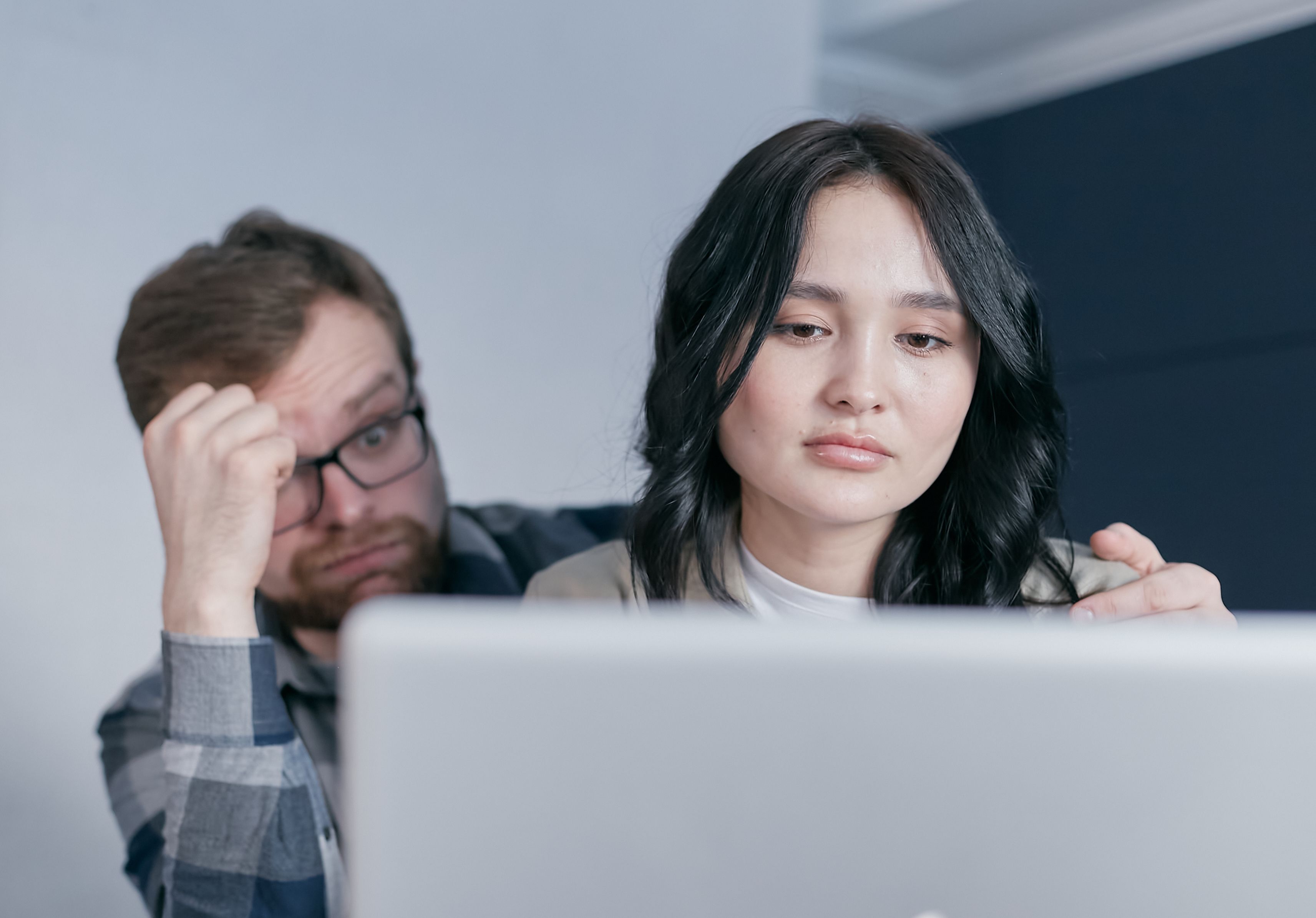 A concerned couple looks at a laptop together. A man wearing glasses rests his hand on his forehead while placing a supportive arm around a woman who appears worried, suggesting financial stress or uncertainty.