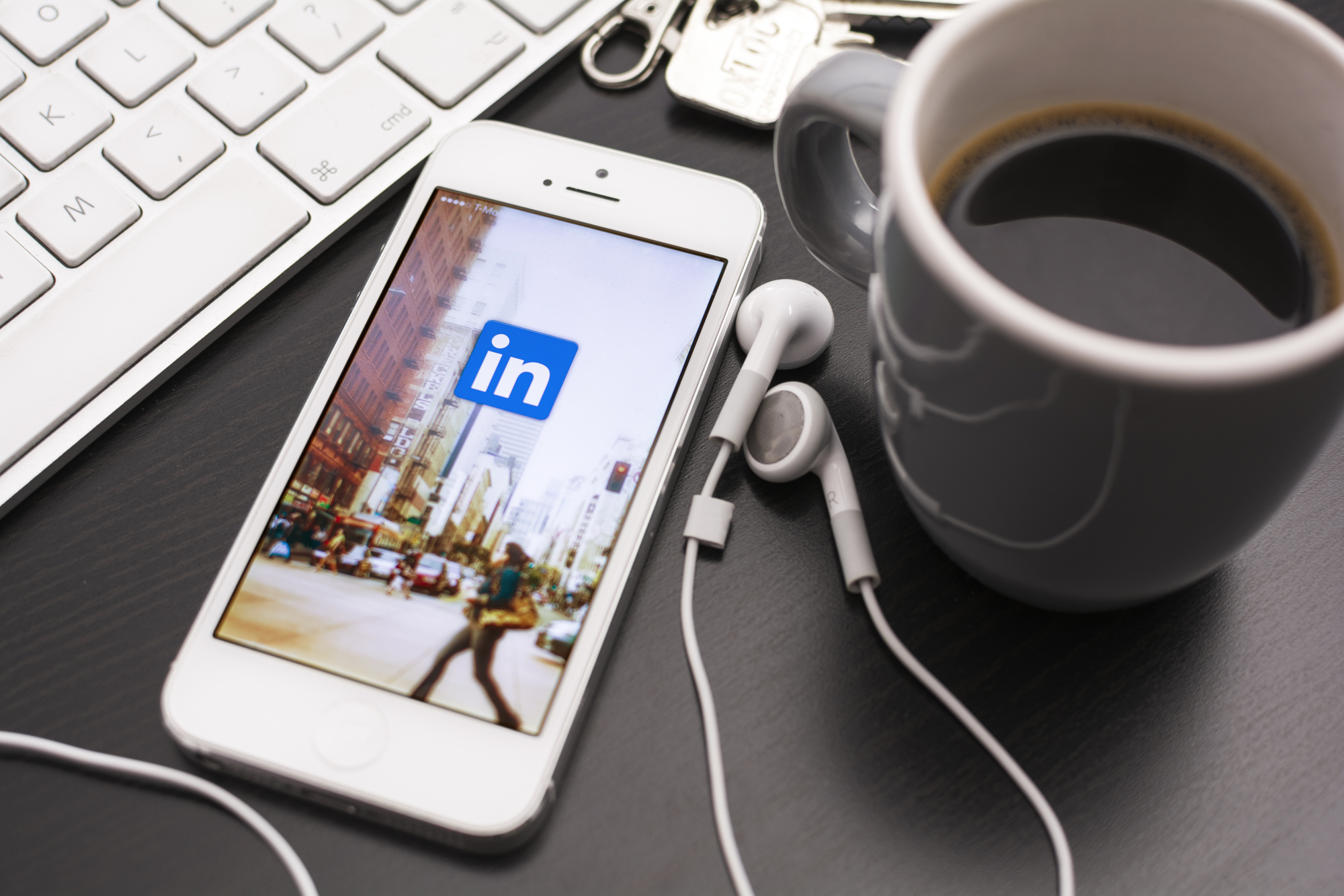 A smartphone displaying the LinkedIn logo sits on a desk beside a computer keyboard, earbuds, keys, and a cup of coffee, representing online professional networking and career development.