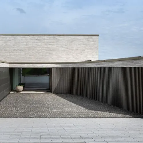 Moderner, minimalistischer Gebäudeeingang mit hellen Ziegelwänden, einem Lattenzaun aus Holz und einer Topfpflanze neben einer Glastür.