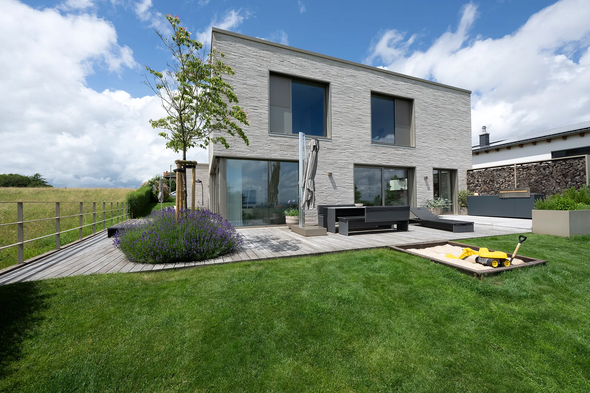 Modernes zweistöckiges Haus mit grauer Steinfassade, großen Fenstern, Holzterrasse mit Gartenmöbeln und einem kleinen Sandkasten mit Spielzeug auf einer grünen Rasenfläche.