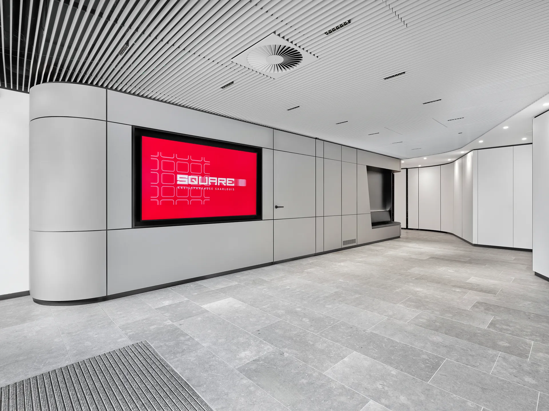 Moderner Innenraum mit grau gefliestem Boden, geschwungenen grauen Wandpaneelen, eingelassener Sitzbank und einem großen roten Digitalbildschirm mit der Aufschrift „SQUARE“.