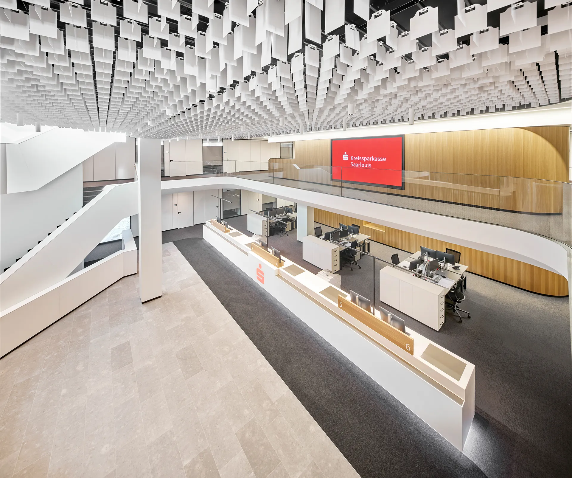 Modernes, geräumiges Bürointerieur mit mehreren Arbeitsplätzen, einem großen roten Digitaldisplay mit der Aufschrift Kreissparkasse Saarlouis und einer Decke mit hängenden weißen rechteckigen Paneelen.