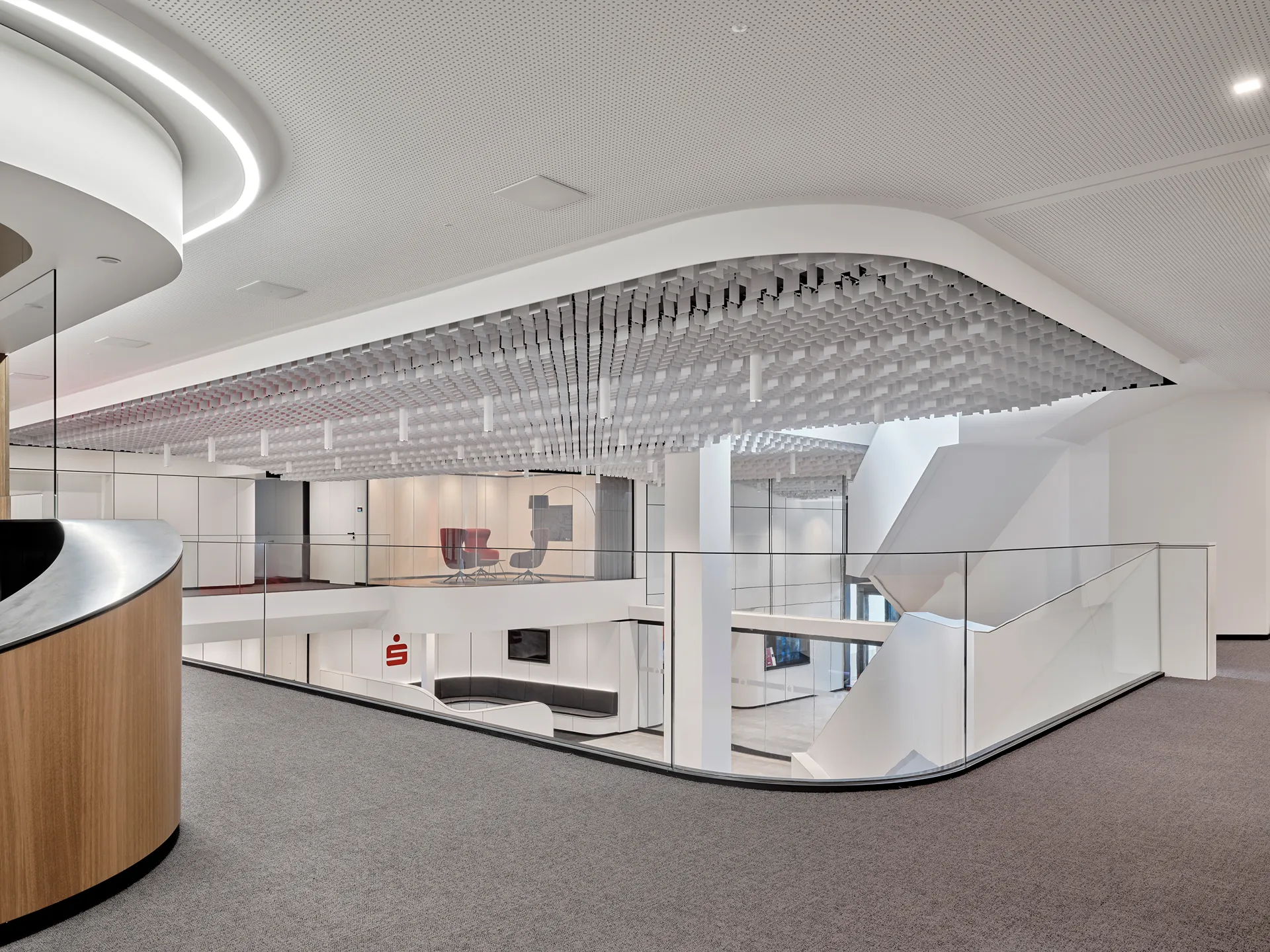 Modernes Bürointerieur mit einer abgehängten Decke aus weißen kubischen Elementen und einem Glasgeländer mit Blick auf ein darunterliegendes Geschoss mit Sitzbereichen.