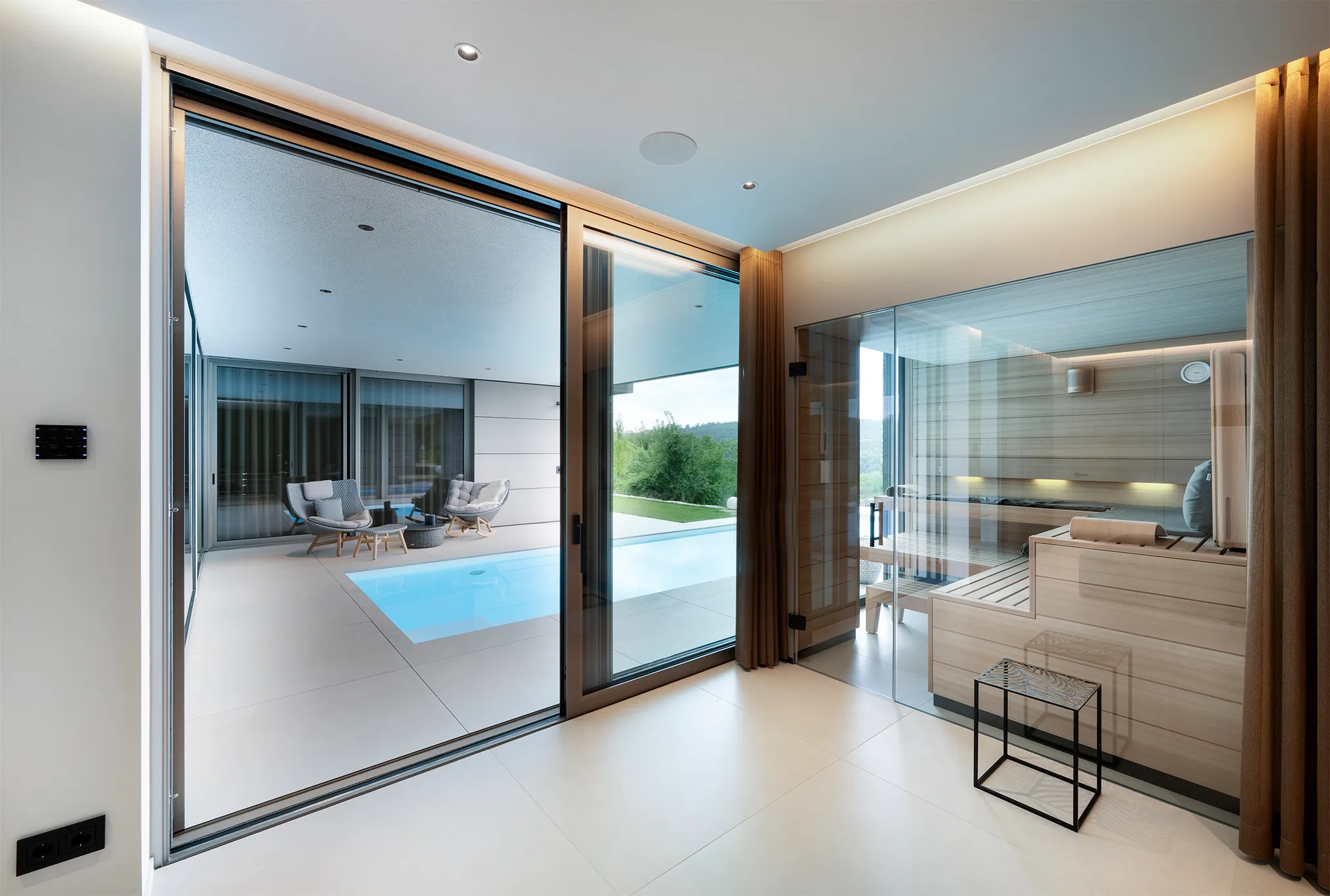 Moderner Innenraum mit einer verglasten Holzsauna auf der rechten Seite und einer Außenterrasse mit kleinem Pool und zwei Liegestühlen.
