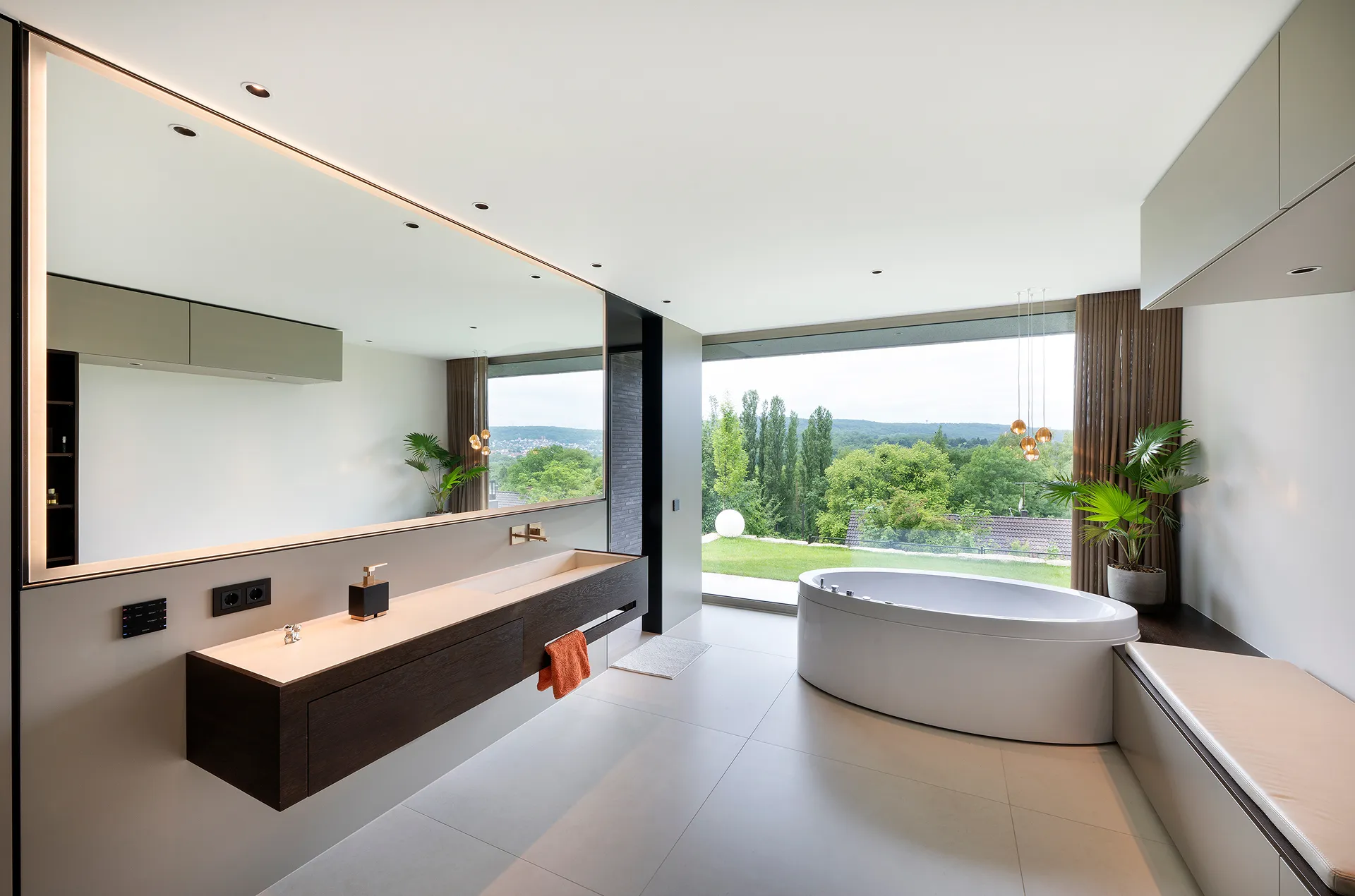 Modernes Badezimmer mit großem Spiegel, schwebendem Waschtisch, ovaler Badewanne und großem Fenster mit Blick auf grüne Bäume und Hügel.