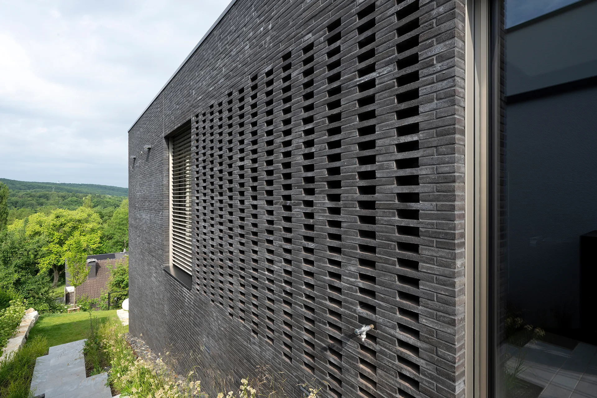 Moderne, dunkle Backsteinmauer mit versetztem, offenem Ziegelmuster und einem Außenwasserhahn, mit Blick auf eine grüne Landschaft.