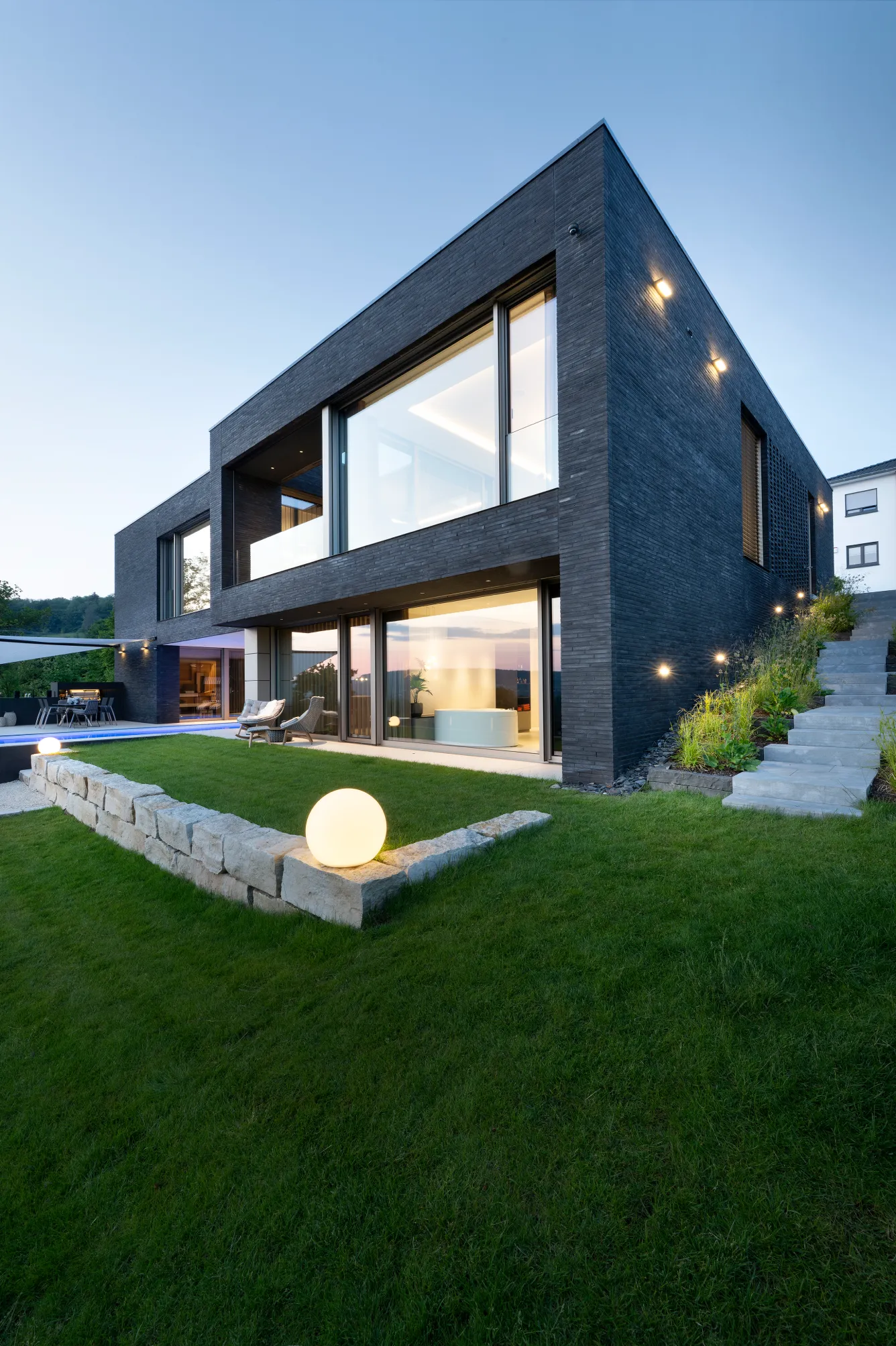 Modernes zweistöckiges Haus mit dunkler Backsteinfassade, großen Fenstern, Sitzgelegenheiten im Freien, leuchtenden kugelförmigen Gartenleuchten und einem Rasen.
