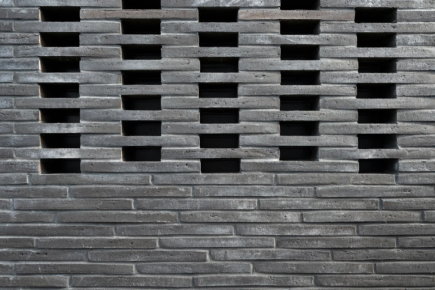 Graue Backsteinmauer mit versetzt angeordneten rechteckigen Öffnungen in horizontalen Reihen.