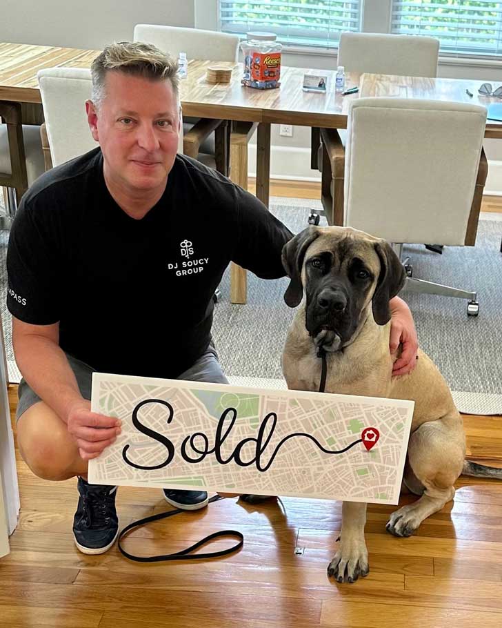 A man kneels on the floor next to a large boxer dog, holding up a sign that says "sold"