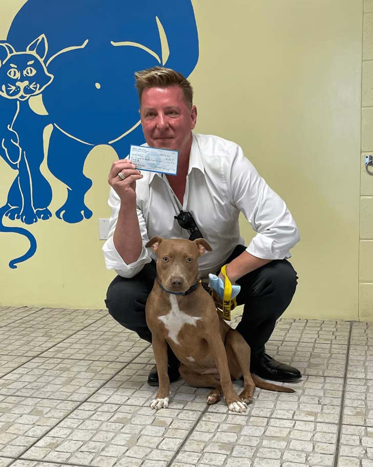 A man in an animal shelter kneels behind a puppy and holds up a check