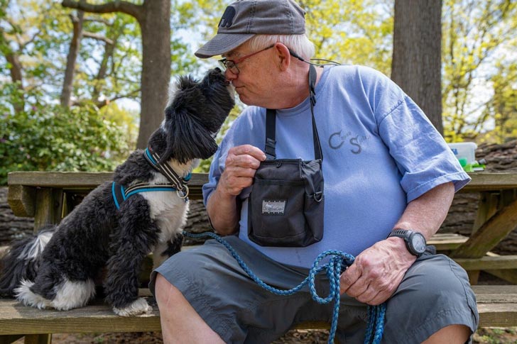 An older gentleman sits on a wooden park bench outdoors. He leans in to his dog, a black and white Tibetan terrier, who lifts his nose to sniff the owner.