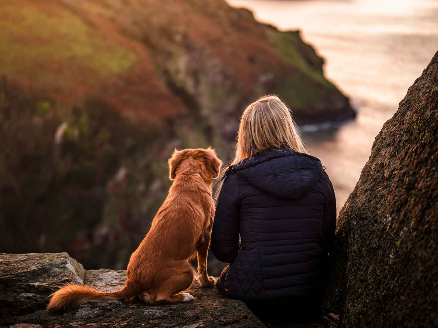 A woman in a black coat sits next to a fluffy brown dog, looking off into a canyon