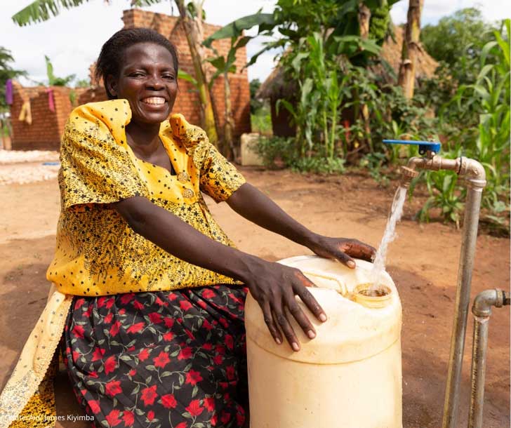 A woman gets fresh water from a spiget in the ground, smiling