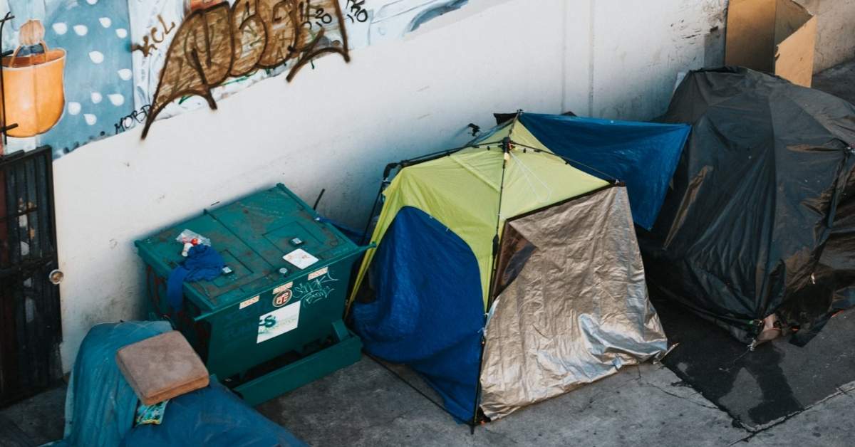 An assortment of tents on a sidewalk against a graffitied city wall. 
