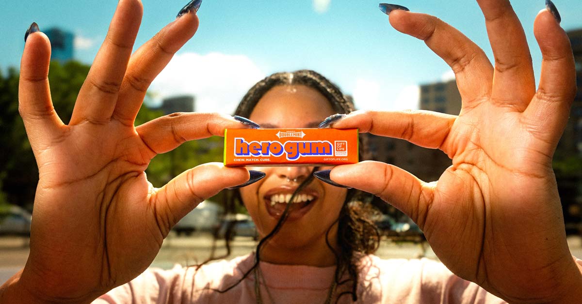 A person holds up a pack of gum in front of their face so it's covering their eyes, but revealing a smiling mouth. The gum is wrapped in orange packaging with blue text that reads "hero gum."
