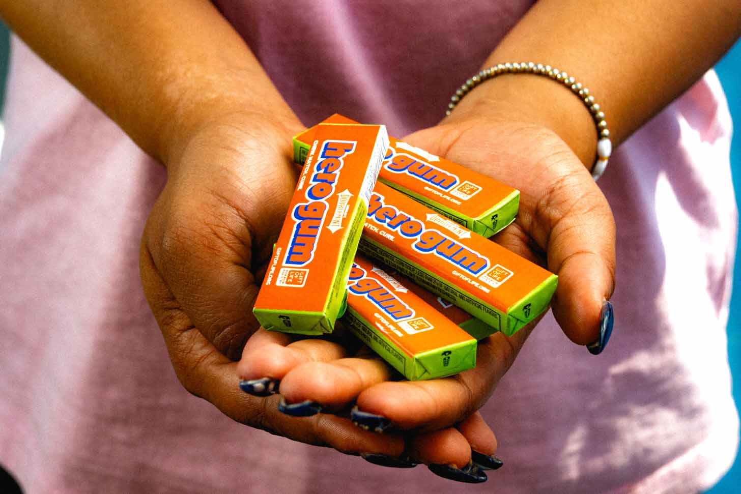 A person holds up a pack of gum in front of their face so it's covering their eyes, but revealing a smiling mouth. The gum is wrapped in orange packaging with blue text that reads "hero gum."