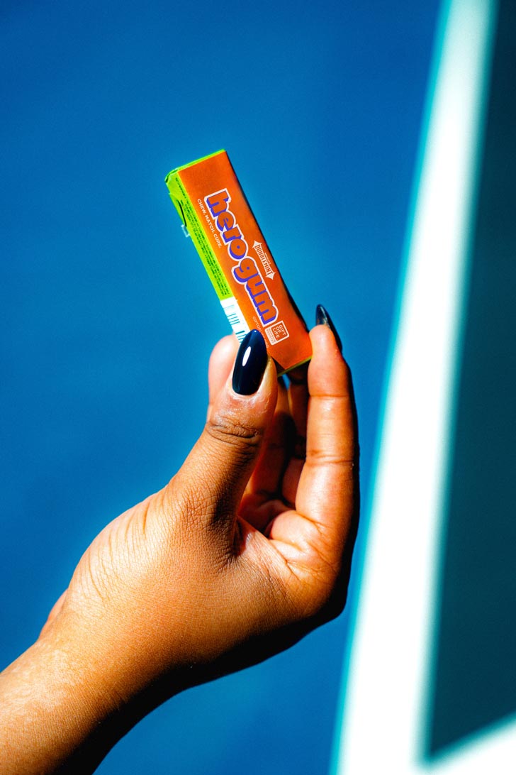 A hand holds a stick of gum against a blue backdrop. It is packaged in an orange casing with blue text that reads: "hero gum."
