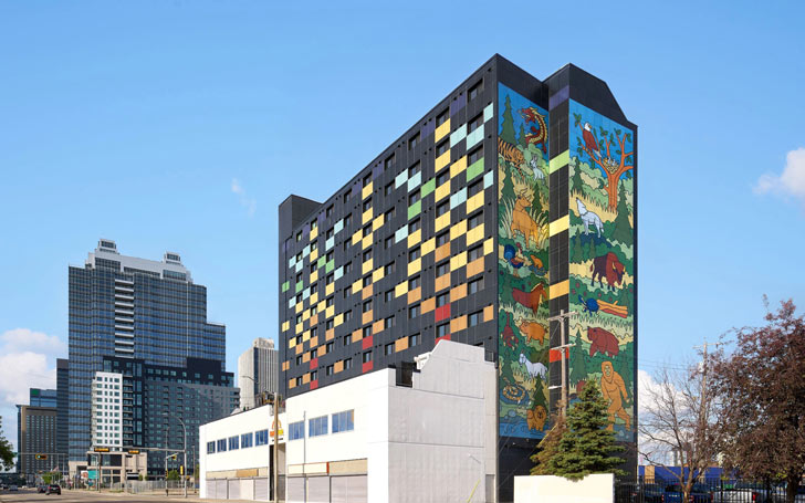 The SunRise Building in Alberta, Canada against a blue, nearly cloudless sky. The exterior of the 12-story residential building is covered in colorful photovoltaic solar panels, forming a mural of animals in nature