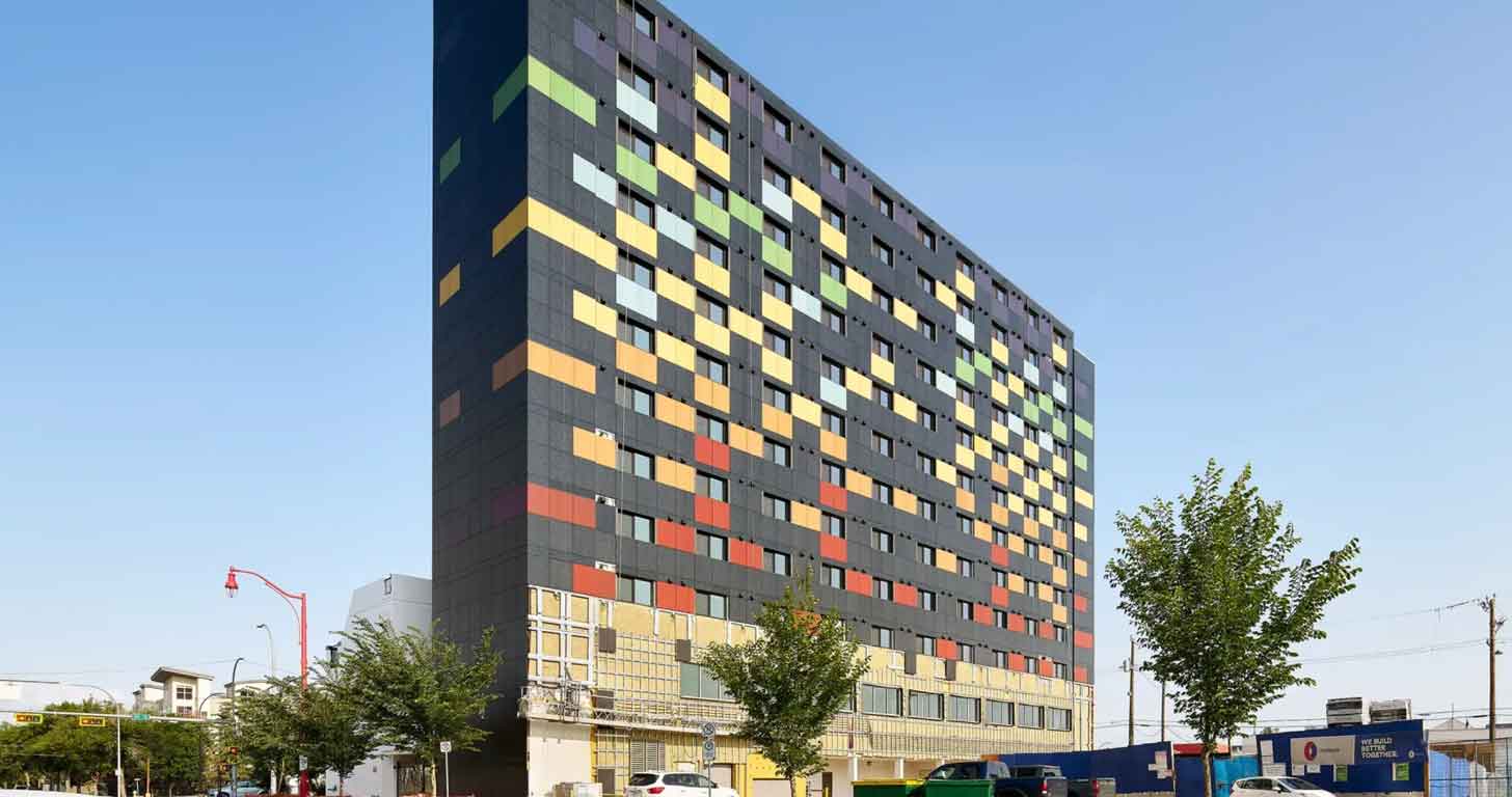 A 12-story building against a blue sky, with a black exterior, and colorful solar panels lining the facade, in red, orange, yellow, blue, green, and purple