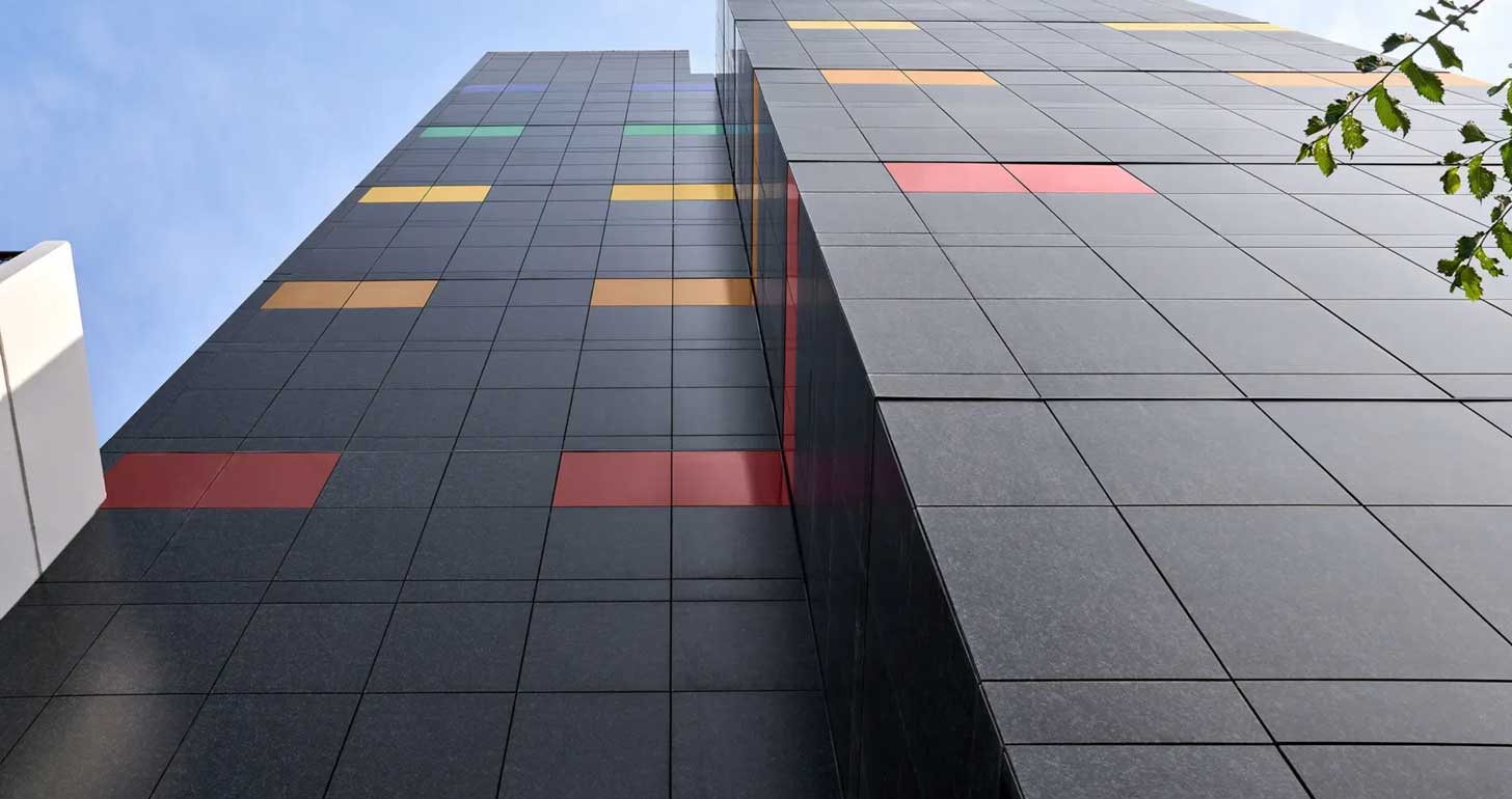 A bottom-up view of a building covered in black, red, orange, yellow, green, and blue solar panels