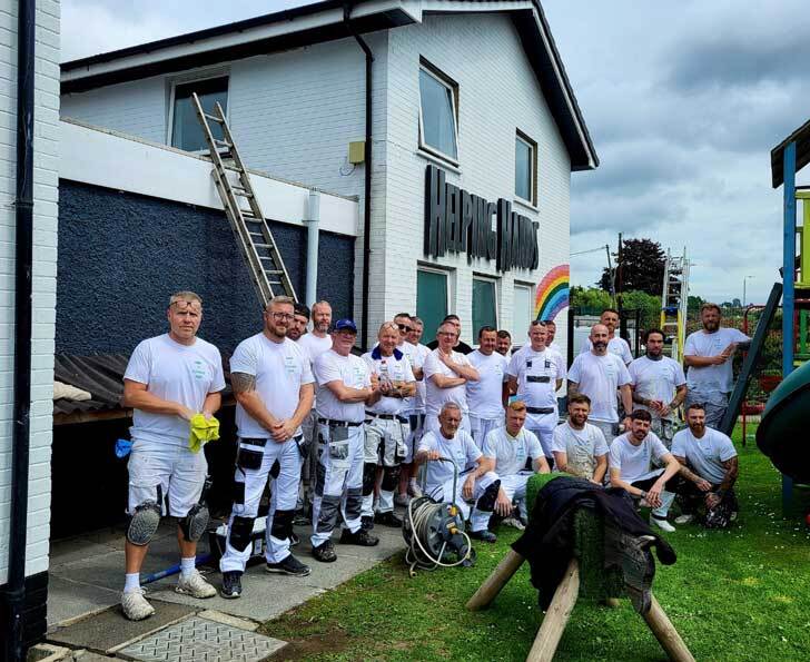 A crowd of men and women in painters uniforms stand infront of a freshly painted white building with rainbow decor. 