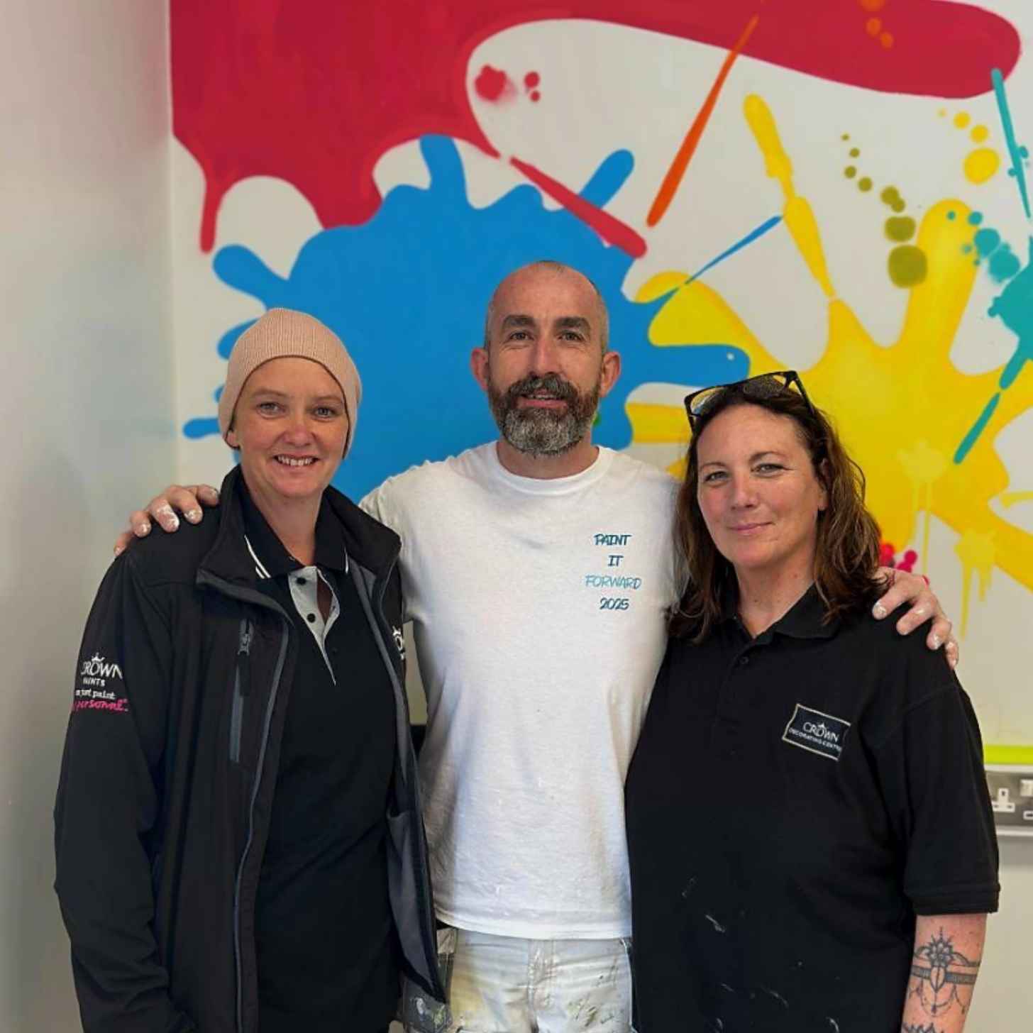 Gerard Donaghy, a white Irish man, smiles as he puts his hands around a woman on his left and right. The wall behind them is full of bright splashes of color. 