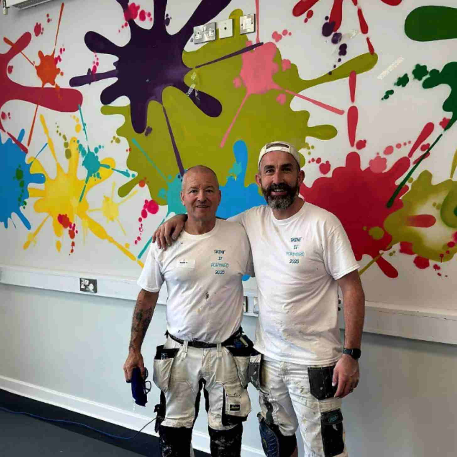 Gerard Donaghy, a white Irish man, smiles as he puts his hands around another white Irish man who volunteered to help paint. The wall behind them is full of bright splashes of color. 